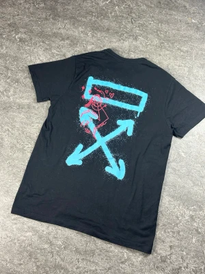 Svart Off-White t-shirt med tryck - Cool svart t-shirt från Off-White med stort grafiskt tryck på ryggen i blått och rosa. Trycket har pilar och en tecknad figur med hjärtan. Klassisk passform och rund hals. Perfekt för dig som vill sticka ut med streetwear-vibe.