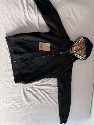 Svart hoodie från Burberry med dragkedja - Säljer en svart hoodie från Burberry med dragkedja och klassiskt rutigt foder i huvan. Hoodien har fickor framtill, ribbade muddar och guldfärgade detaljer på snöret. Perfekt för dig som vill ha en stilren och lyxig look. Den är helt oanvänd. Står att den är XL men det är för barn så den passar även vuxen med storlek M/S mycket bra också.