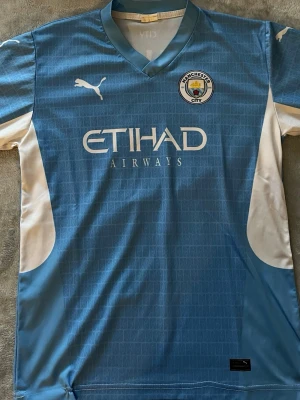 Manchester City matchtröja Mahrez 26 - Snygg ljusblå Manchester City matchtröja från Puma med Mahrez och nummer 26 på ryggen. Tröjan har klubbmärke och Etihad Airways-tryck på bröstet, vita detaljer på sidorna och korta ärmar. Perfekt för fotbollsfans som vill visa sitt stöd på matchdag.