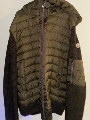 Olivgrön pufferjacka från Moncler - Snygg olivgrön pufferjacka från Moncler med stickade mörkbruna ärmar och huva. Jackan har dragkedja framtill, två fickor med knapp och en extra ficka på ärmen. Moncler-logga på vänster ärm. Perfekt för kalla dagar och riktigt trendig look.