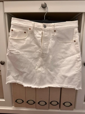 Vit levis jeanskjol med rå kant - Säljer en vit kort jeanskjol med fem fickor och rå, fransig kant nertill. Kjolen har slitna detaljer framtill för en cool, avslappnad look. Passar perfekt till sneakers eller boots för en trendig outfit.