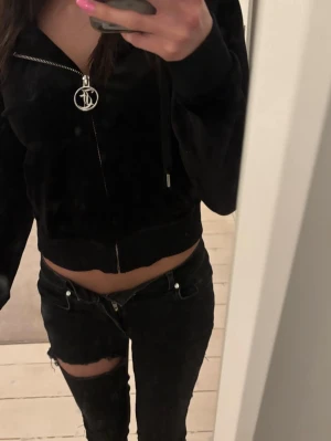 Svart velour hoodie från Juicy Couture - Säljer en svart croppad velour hoodie från Juicy Couture med dragkedja och ikonisk JC-logga på zip-pullern. Hoodien har huva med snören och bred mudd i midjan. Perfekt för en chill och trendig look.