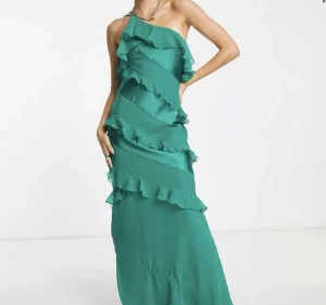 Grön långklänning med volangdetaljer - Säljer en snygg grön långklänning med volangdetaljer och oneshoulder-design. Klänningen är använd en gång under min bal och har inga  fläckar🩷. Storleken är 34 men passar mig som har storlek 36 bra!! 