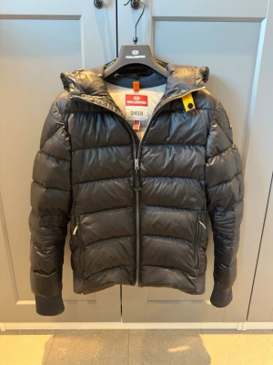 Svart pufferjacka från Parajumpers - Snygg svart pufferjacka från Parajumpers med huva och dragkedja framtill. Samt två fickor med dragkedja. Tillverkad i glansigt material och har en gul detalj vid halsen. Passar de som har stl S och på gränsen till M . Tyvärr har den fått ett litet hål på ärmen och lite skrap, men inget som förstör jackans snygghet