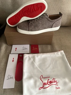 Gråa sneakers från Christian Louboutin - Snygga gråa sneakers från Christian Louboutin i mocka med klassisk röd sula och vita kanter. Skorna har en stilren design med matchande grå snörning och diskreta sömmar. Perfekta för dig som vill ha något exklusivt och trendigt i garderoben.