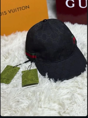Svart keps från Gucci med logga - Snygg svart keps från Gucci med diskret GG-monogram över hela tyget och klassiska gröna och röda ränder på sidan. Justerbar rem baktill i läder och metallspänne. Tillverkad i bomullsmix och har märkets etikett på insidan.