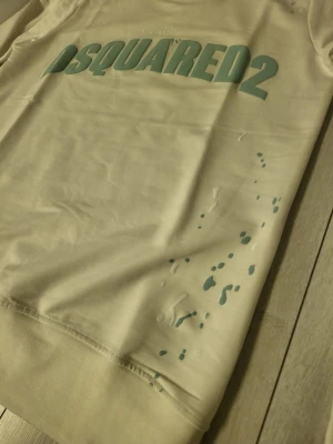 Vit Dsquared2 t-shirt med tryck - Säljer en vit tröja från Dsquared2 med stort grönt logotyp-tryck på bröstet. Klassisk rund hals . Tillverkad i mjuk bomull som känns skön mot huden. Perfekt för dig som gillar streetwear och vill sticka ut med ett tydligt märke.. riktigt bra kvalitet 