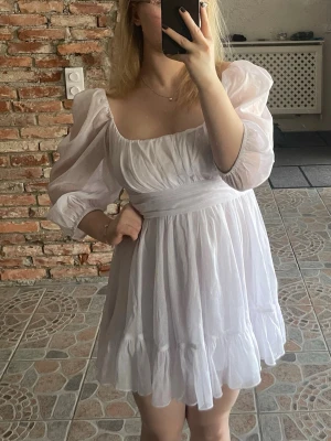 Vit kort skimrande barmaidklänning med puffärmar och knyt - Supersöt vit kort så kallad barmaid klänning med lätt skimmer invävt puffiga trekvartsärmar och stor rosett i ryggen. Klänningen har volangkant nertill och är gjord i ett lätt, luftigt tyg som passar perfekt till sommaren. Använd på student 🥰