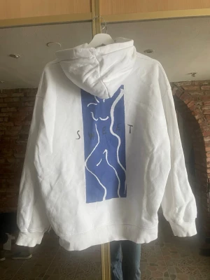 Vit hoodie med blått tryck på ryggen - Säljer en oversized vit hoodie med huva och känguruficka. På ryggen finns ett stort blått konstnärligt tryck med en stiliserad kropp och texten 'SWEET'. Hoodien är i mjukt material, perfekt för en chill och avslappnad stil. Finns dessvärre en fläck på framsidan se sista bilden 🌸