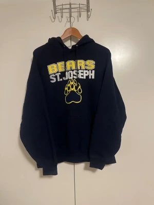 Vintage Marinblå hoodie  - Snygg marinblå Vintage hoodie med trycket 'BEARS ST. JOSEPH' i gult och vitt samt en gul björntass på bröstet. Klassisk huva med dragsko och stor magficka framtill. Perfekt för dig som gillar college-stil och vill ha en bekväm och avslappnad look.