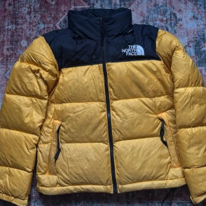 Gul och svart dunjacka The North Face retro inspirerad - Gul och svart dunjacka från The North Face med hög krage, dragkedja framtill och två sidofickor med dragkedja. Jackan har broderad logga på bröstet och justerbar dragsko vid midjan samt gömd luva. Tror den är retro inspirerad och har tjänat mig väl under tiden jag använt den. Inga defekter. Pris kan diskuteras!!!
