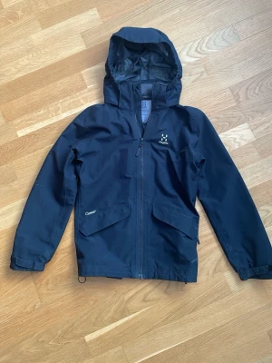 Svart vindjacka från Haglöfs - Snygg svart vindjacka från Haglöfs med huva och dragkedja framtill. Jackan har två stora fickor med lock och justerbara ärmslut. Perfekt för blåsiga dagar och outdoor-äventyr.