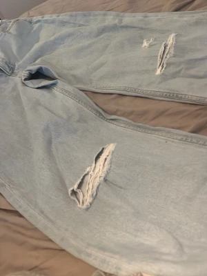 Ljusa ripped jeans från H&M - Säljer ett par ljusblå jeans från H&M med hög midja och justerbar midja. Jeansen har bred passform och slitna detaljer med stora hål på benen för en trendig look. Perfekta för dig som gillar streetstyle och vill ha ett par riktigt snygga ripped jeans.