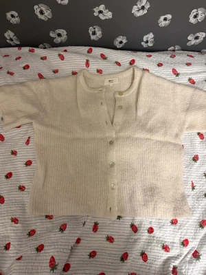 Krämvit kofta från Gina Tricot - Mysig krämvit kofta från Gina Tricot i ullblandning. Koftan har korta ärmar, ribbad struktur och knappar framtill. Perfekt att slänga över en topp för en chill look. Enkel och stilren design som passar till många outfits.