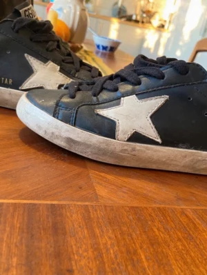 Svarta Golden Goose sneakers med vit stjärna - Coola svarta sneakers från Golden Goose med vit stjärna på sidan och vita sulor. Skorna har svarta skosnören och GGDB-logga på tungan. Tillverkade i läder och har en klassisk låg modell som är enkel att matcha med olika outfits. Det är 35 men passar 36-37.