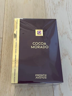 Cocoa Morado Eau de Parfum 100ml - Cocoa Morado från French Avenue är en parfym i en elegant mörklila och guld kartong. Flaskan innehåller 100 ml Eau de Parfum. Förpackningen har en lyxig känsla med gulddetaljer och en lila dekorsten på framsidan. Perfekt för dig som gillar exklusiva dofter. Den är helt oöppnad.