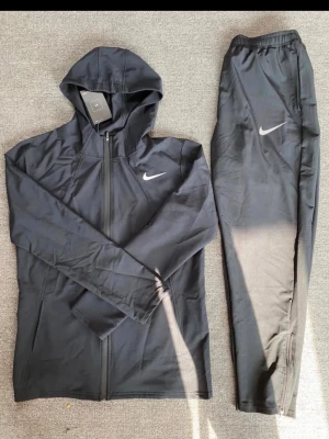 Svart Nike träningsset med huva - Säljer ett svart träningsset från Nike med zip-hoodie och matchande byxor. Setet har vita Nike-loggor på både jackan och byxorna. Hoodien har dragkedja och byxorna har resår i midjan. Perfekt för gymmet eller löpning.