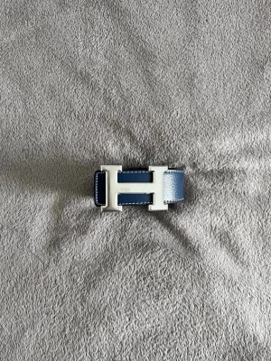 Herme’s bälte  - Säljer ett stilrent blått skärp med ett ikoniskt H-formad spänne i silver. Skärpet är tillverkat i skinn och har en slät, elegant design som passar perfekt till jeans eller kostymbyxor. Ett riktigt statement-accessoar för dig som gillar klassiska detaljer.