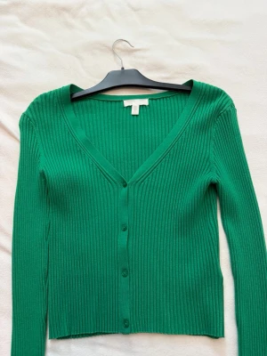 Grön ribbad kofta från H&M - Snygg grön ribbad kofta från H&M med v-ringning och knappar framtill. Koftan har långa ärmar och är tillverkad i ett stretchigt material som sitter skönt på kroppen. Perfekt att slänga över en t-shirt eller topp för en cool look.