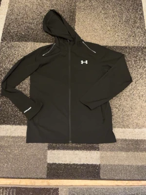 Under Armour Storm-jacka - Svart windbreaker från Under Armour med hel dragkedja och huva. Jackan har reflektiva detaljer på axlarna, logga på bröstet och stort Under Armour-tryck på ryggen. Tillverkad i lätt syntetmaterial, perfekt för träning eller chill.