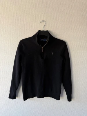 Svart halvzip tröja från Polo Ralph Lauren - Svart långärmad tröja från Polo Ralph Lauren med halvzip och hög krage. Diskret broderad logga på bröstet. Tröjan är i mjukt material och har en klassisk, stilren design som passar perfekt till jeans eller chinos. Har en defekt vid höger armbåge.