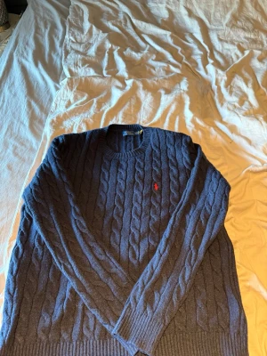 Mörkblå kabelstickad tröja Polo Ralph Lauren - Säljer en mörkblå kabelstickad tröja från Polo Ralph Lauren med rund hals och klassisk röd logga broderad på bröstet. Tröjan har långa ärmar och ribbade muddar vid ärmslut och nederkant. Perfekt för dig som gillar stilrena och tidlösa plagg.