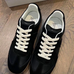 Maison Margiela Gats storlek 45 - Svarta sneakers från Maison Margiela med vita snören och beige sula. Skorna har en klassisk siluett med paneler i både mocka och läder, samt vit insida. Perfekta för en clean och stilren look.