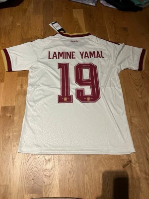 Spanien Lamine Yamal matchtröja - Säljer en vit Spanien fotbollströja från Adidas med vinröda detaljer och nummer 19 samt Lamine Yamal på ryggen. Tröjan har korta ärmar, guldfärgade inslag vid krage och ärmslut samt officiellt landslagsmärke på bröstet. Tillverkad i lätt och ventilerande material.