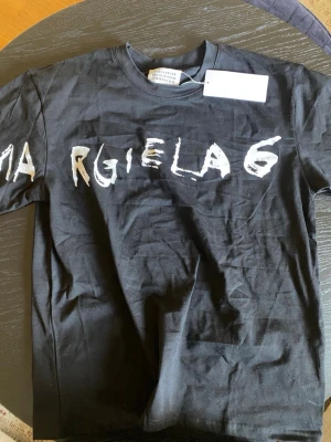 Svart Maison Margiela t-shirt - Cool svart t-shirt från Maison Margiela med vit, handmålad text över bröstet. Klassisk rund halsringning och korta ärmar. Perfekt för dig som gillar streetwear och vill sticka ut med en unik design. Materialet är mjuk bomull.
