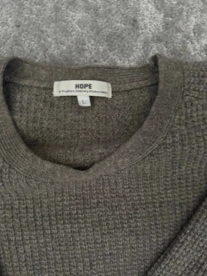 Grå stickad tröja från HOPE - En stilren grå stickad tröja från HOPE med rund halsringning och diskret ribbstickad struktur. Tröjan har långa ärmar och breda muddar vid ärmslut och nederkant. Perfekt för lager-på-lager och enkel att matcha med jeans eller chinos.
