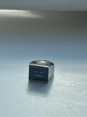 Silverfärgad ring Acne Studios Face - Cool silverfärgad moodring från Acne Studios med deras ikoniska Face-motiv på en fyrkantig platta. Ringen har en modern och minimalistisk design som sticker ut och passar perfekt för dig som gillar statement-smycken. Tillverkad i metall med svart detalj på framsidan.