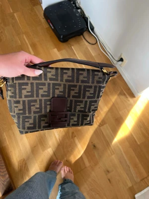 brun handväska - Snygg brun handväska  i mörkbrunt och beige. Väskan har ett lock med brunt spänne och axelrem i läder. Guldiga metalldetaljer och rymligt innerfack. Perfekt för dig som gillar lyxiga accessoarer och tidlös design.