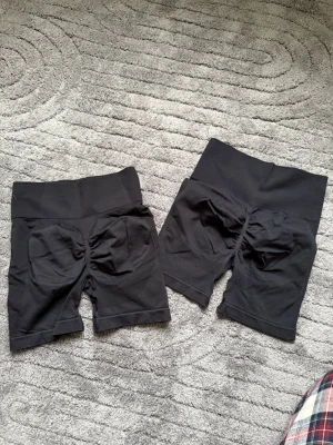 Svarta träningsshorts - Två par svarta högmidjade träningsshorts i stretchigt syntetmaterial. De har bred resår i midjan och rynkade detaljer bak för en snygg form. Perfekta för gymmet eller andra sportaktiviteter där du vill ha en tight och bekväm passform. Båda är oanvända och storlek s