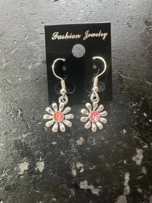 Blommiga örhängen med rosa sten - Säljer ett par söta örhängen i form av blommor med silverfärgade kronblad och en rosa glittrande sten i mitten. Örhängena har krokfäste och ger en lekfull och trendig look. Perfekta för dig som vill addera lite färg och bling till din stil.