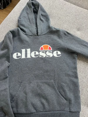 Grå Ellesse hoodie med logga - Säljer en grå hoodie från Ellesse med stor vit och orange logga på bröstet. Tröjan har en klassisk huva och en stor magficka framtill. Perfekt för dig som gillar sportig och avslappnad stil.