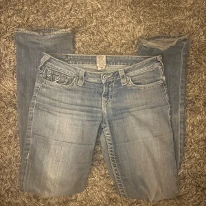 True Religion ljusblå jeans, stl 28 - Ljusblå jeans från True Religion med klassiska kontrastsömmar och snygga fickdetaljer bak. Jeansen har slitningar vid bensluten och en rak passform. Lila kristaller till knappar. Väldigt använda och vintage så slitningar förekommer, fråga om bilder angående.