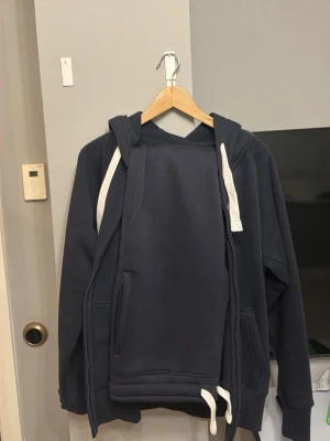 Mörkblå hoodie från Russemerch - Mörkblå hoodie från märket R med vit dragsko och huva. Klassisk design med fickor på framsidan och ribbade muddar. Perfekt för dig som gillar en enkel och stilren look. Materialet är mjukt och bekvämt, passar bra till jeans eller joggers.