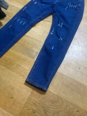 Blå distressed jeans med raka ben - Säljer ett par blå jeans med raka ben och coola slitningar på framsidan. Jeansen har klassisk femficksdesign och är gjorda i denim. Perfekt för dig som gillar en avslappnad och trendig look.