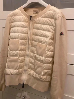 Beige cardiganjacka från Moncler - Snygg och trendig cardigan från Moncler med stickade ärmar och quiltad front. Vit och beige färg. Använd och lite smutsig med sminkfläckar som syns på bilderna, men i övrigt hel och fin utan hål.   Inköpt 2023 hos Monclers butik, Bild på kvitto finns och även tags samt qr-kod till hemsidan intygar äktheten.   Storlek M men passar också S