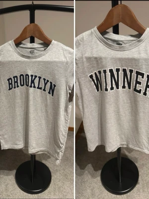 2st Grå t-shirt med tryck från 157 - Säljer två grå t-shirt från 157 med coola tryck på framsidan, 'BROOKLYN' och 'WINNER' i mörkblå text. T-shirten har rund hals och korta ärmar, perfekt för en avslappnad och sportig look. Materialet är mjukt och känns skönt mot huden.