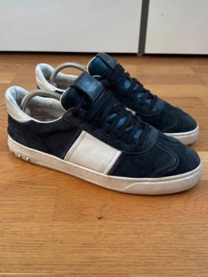 Svarta och vita sneakers från Valentino - Snygga sneakers från Valentino i svart och vitt med klassisk låg profil. Skorna har en vit panel på sidan, svarta skosnören och coola nitar på hälen. Materialet är en mix av mocka och läder, vilket ger en lyxig känsla.