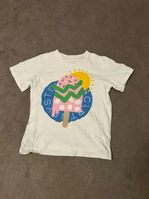 Vit t-shirt Stella McCartney Kids - Vit t-shirt från Stella McCartney Kids med färgglad glass- och sol-illustration framtill. T-shirten är i storlek 8 år. 💛