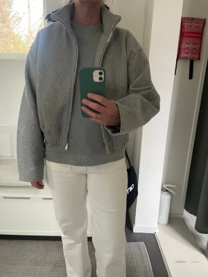 Grå bomberjacka med krage - Säljer en grå bomberjacka med hög krage och dragkedja framtill. Jackan har en boxy passform, elastisk nederkant och är tillverkad i ett mjukt material som liknar ullmix. Perfekt för dig som gillar en clean och stilren look.