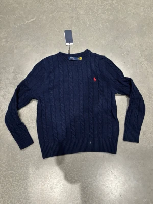 Ralph Lauren tröja - Kabelstickad mörkblå tröja från Polo Ralph Lauren med rund halsringning och röd broderad logga på bröstet. Tröjan har ribbade muddar vid ärmslut och nederkant, vilket ger en klassisk och stilren look. Perfekt för dig som gillar tidlös design.