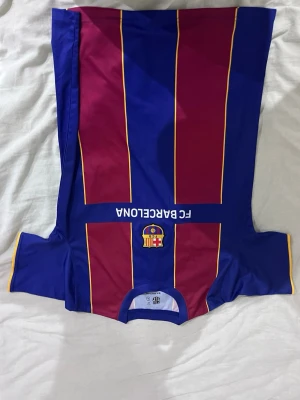 FC Barcelona matchtröja blå/röd - Snygg FC Barcelona matchtröja i blått och vinrött med gula detaljer. Tröjan har klubbens logga på bröstet och texten FC BARCELONA. Perfekt för dig som älskar fotboll och vill visa ditt stöd för laget.