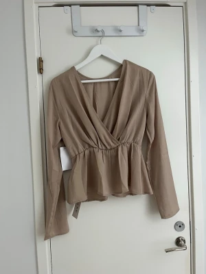 Beige omlottblus från Nakd - Snygg beige omlottblus med djup v-ringning. Blusen har långa ärmar och är tillverkad i ett lätt och flowigt material som ger en skön känsla. Perfekt för både vardag och utekväll. Blusen är från Nakd och är i storlek 36. Den är aldrig använd och har lappen kvar! Toppen går även att ha på sig på båda håll🤩