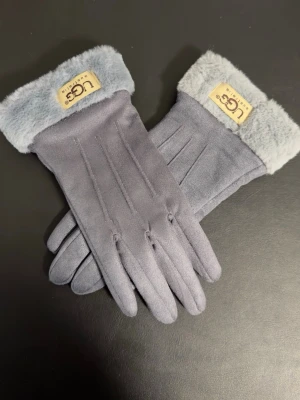 Ugg Grå mockahandskar - Snygga grå mockahandskar från UGG med mjuk pälsdetalj vid handleden och diskret logotyp. Handskarna har en stilren design med tre sömmar på ovansidan och är perfekta för att hålla händerna varma under kalla dagar.