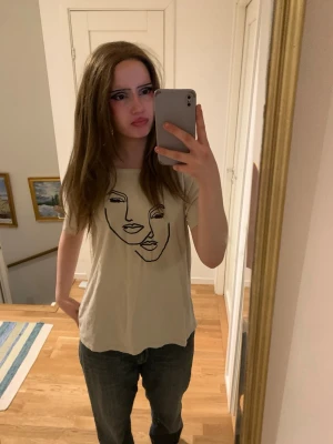 Beige t-shirt med ansiktsmotiv - Säljer en beige t-shirt med coolt svart ansiktsmotiv på framsidan. T-shirten har en avslappnad passform och korta ärmar, perfekt för dig som gillar stilrena och konstnärliga prints. Materialet känns mjukt och skönt mot huden.