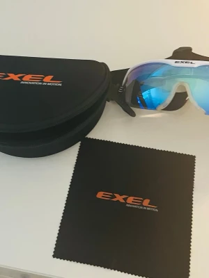 Blå sportiga solglasögon från Exel - Säljer ett par sportiga solglasögon från Exel med vit båge och spegelblått lila glas. Perfekta för dig som vill ha en cool och funktionell look till utomhusaktiviteter. Endast använda 2-3 gånger. Ny pris runt 800 kr