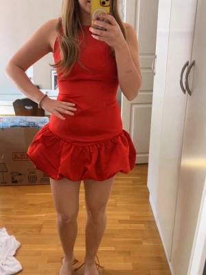 Röd kort klänning med volang - Säljer en röd kort klänning med volang nedtill. Klänningen är ärmlös och har en figurnära passform upptill med en utställd kjol. Perfekt för dig som vill sticka ut med en färgstark och trendig look.